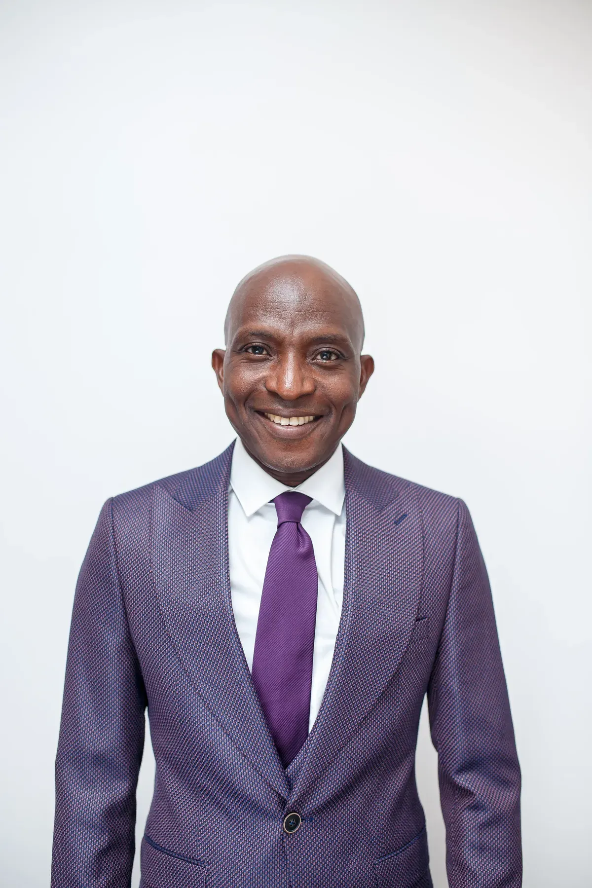Portrait of Richard Meseko in a suit and purple tie against a white wall.